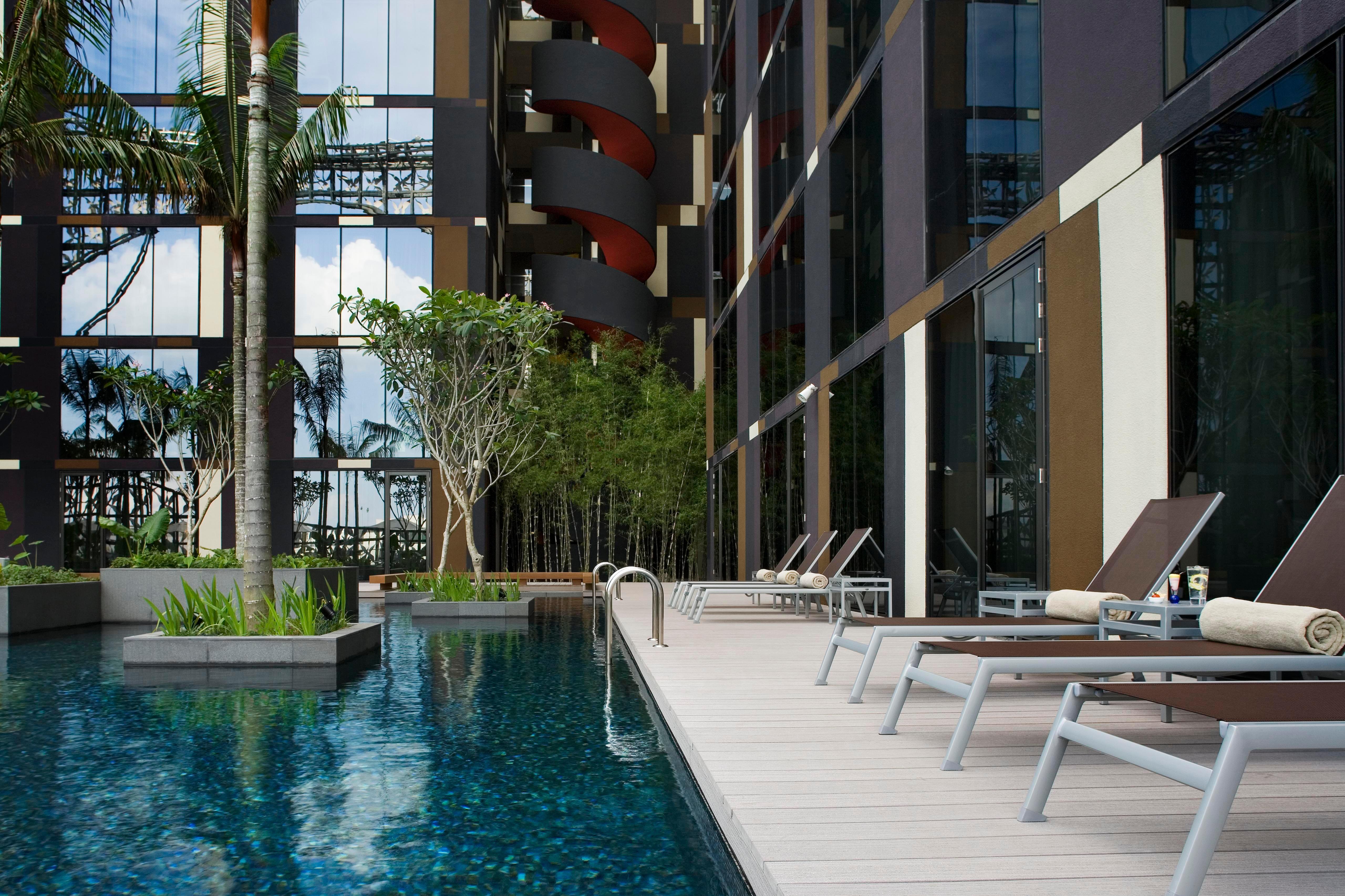 Crowne Plaza Changi Airport By Ihg Сінгапур Зручності фото The photo shows a modern swimming pool area adjacent to a building. The pool is surrounded by sleek wooden decking and features a clear blue water surface. There are several lounge chairs positioned around the pool for relaxation. Lush greenery, incl