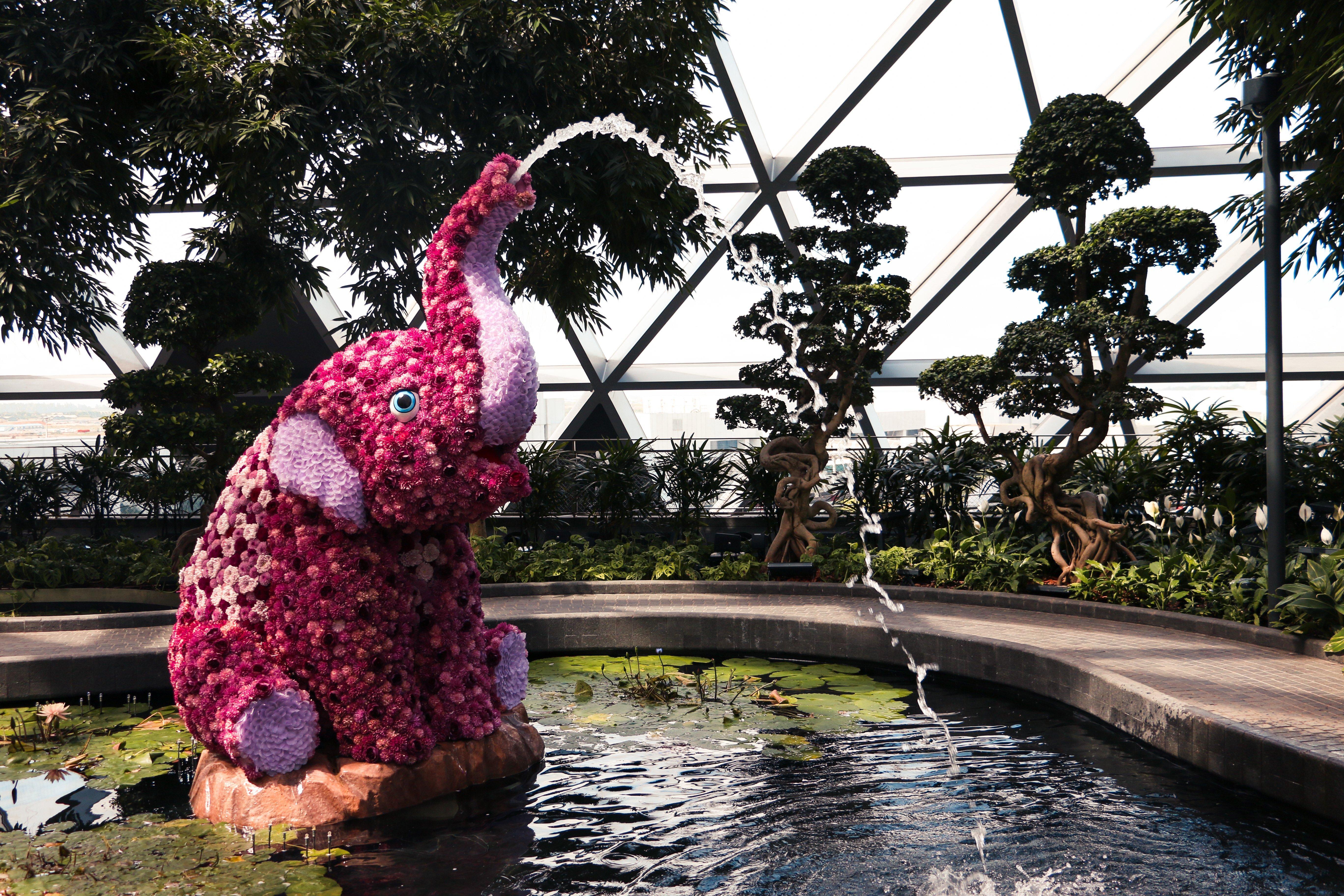 Crowne Plaza Changi Airport By Ihg Сінгапур Екстер'єр фото The photo depicts a decorative elephant sculpture made of flowers, positioned near a pond. The elephant is sitting on its hind legs, with its trunk raised high, creating a water fountain effect. Surrounding the elephant are lush green plants and tree