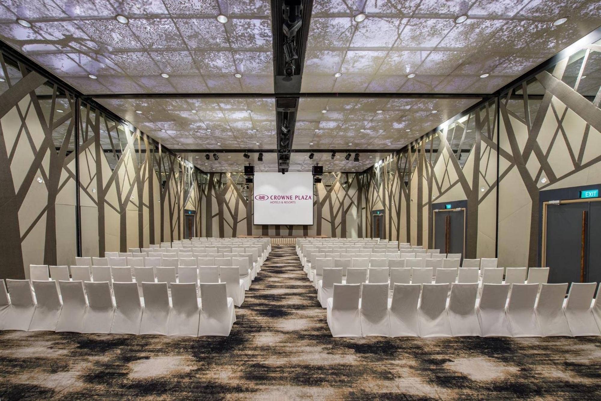 Crowne Plaza Changi Airport By Ihg Сінгапур Екстер'єр фото The photo shows a modern conference or event space. The room features rows of white chairs arranged in a theater-style layout, facing a large screen with a simple logo. The ceiling is designed to resemble a natural setting, with images that suggest t