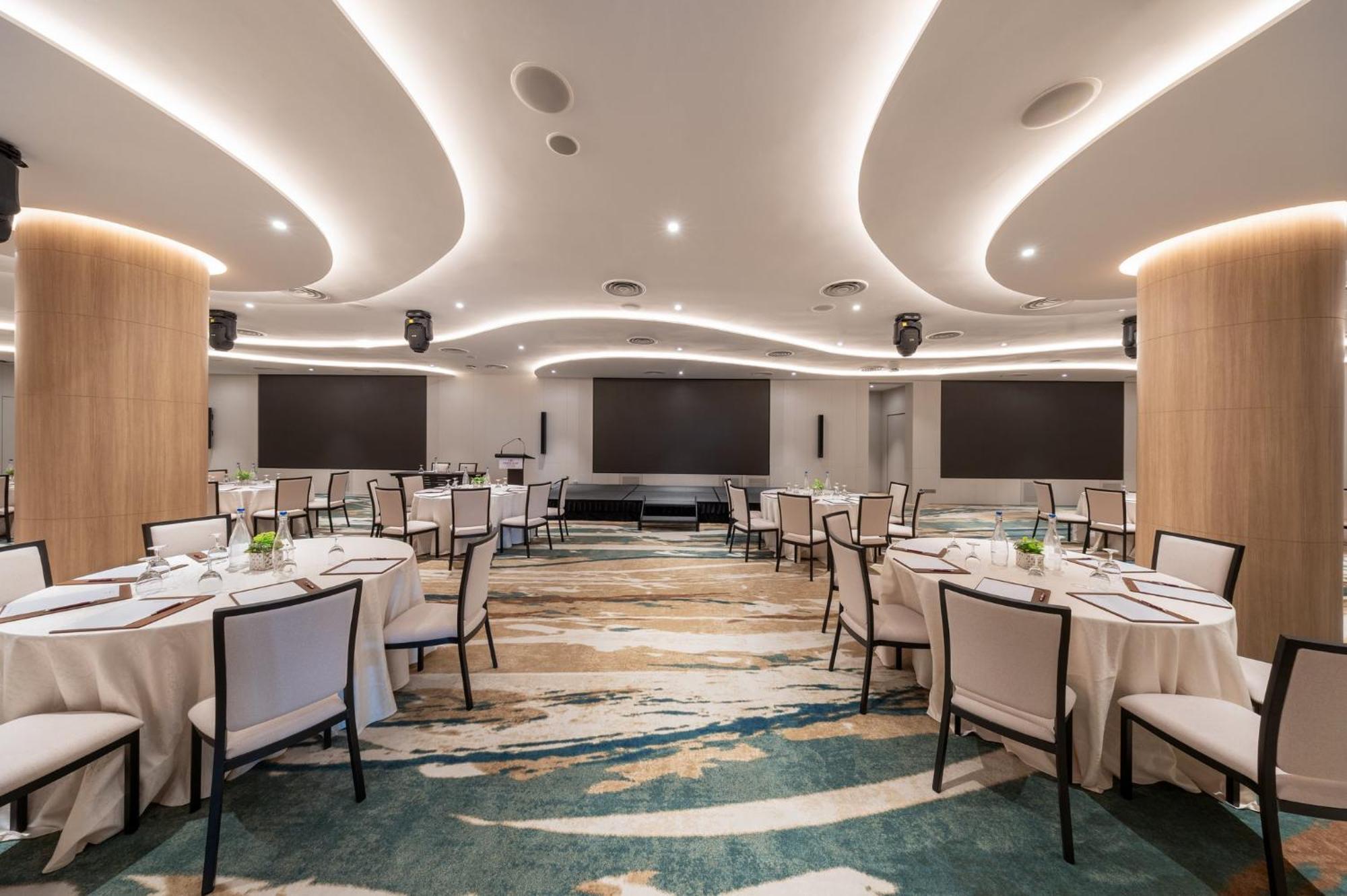 Crowne Plaza Changi Airport By Ihg Сінгапур Екстер'єр фото The photo shows a modern banquet or conference room. The room features elegantly arranged round tables with white tablecloths and chairs. The flooring has an artistic design that combines various shades of blue and beige. The ceiling is accentuated w