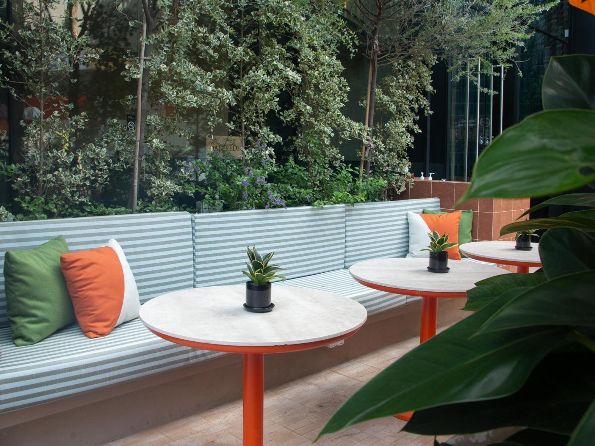 Crowne Plaza Changi Airport By Ihg Сінгапур Екстер'єр фото The photo shows a modern outdoor seating area. There is a long bench with a corrugated metal backrest, decorated with colorful cushions in green and orange. In front of the bench, there are circular tables with white marble tops and orange legs. Each