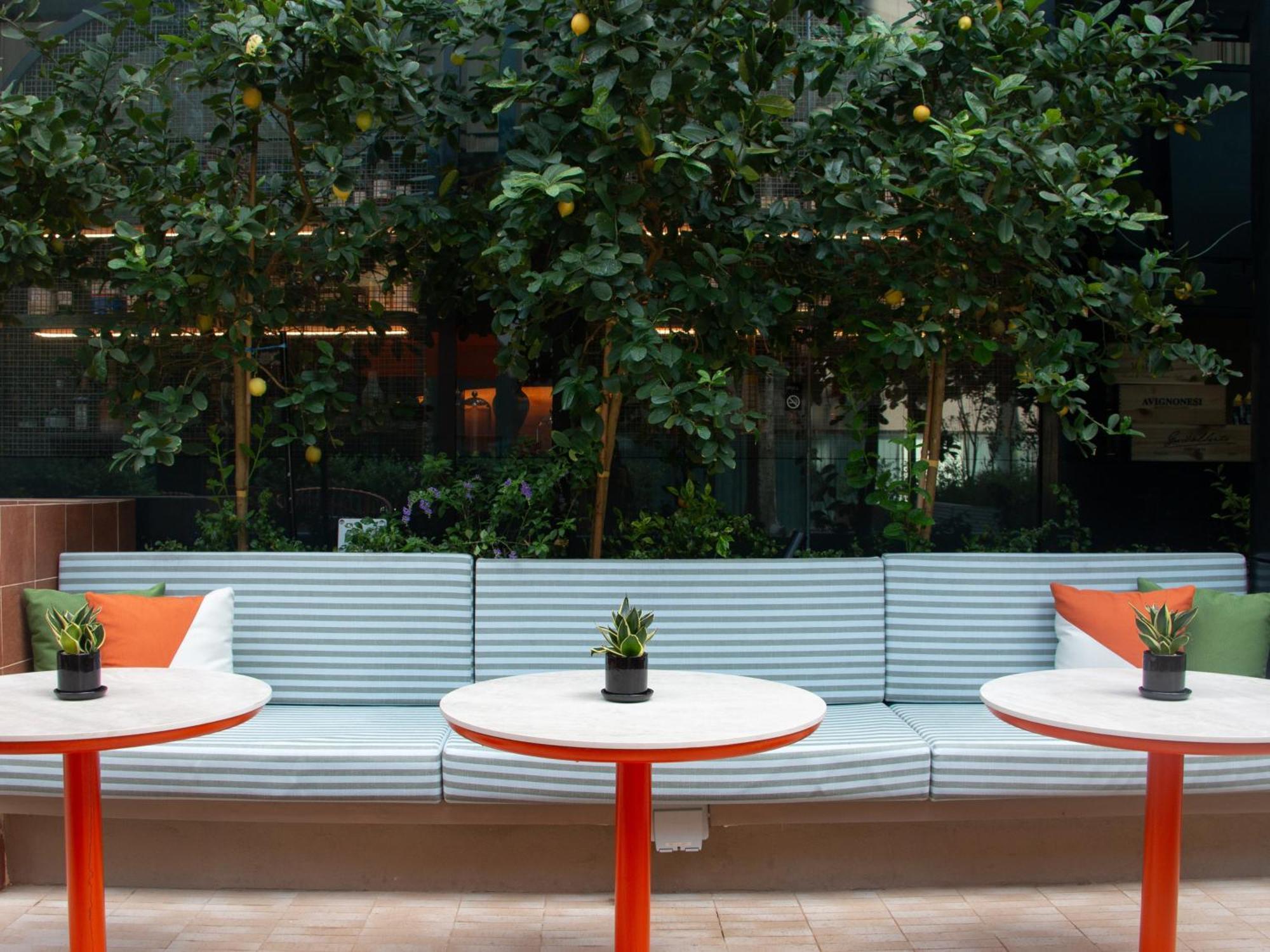 Crowne Plaza Changi Airport By Ihg Сінгапур Екстер'єр фото The photo shows a cozy seating area featuring a long, light blue bench with colorful cushions in orange and green. In front of the bench, there are three round tables with bright red bases and small plants in black pots on each table. The background 
