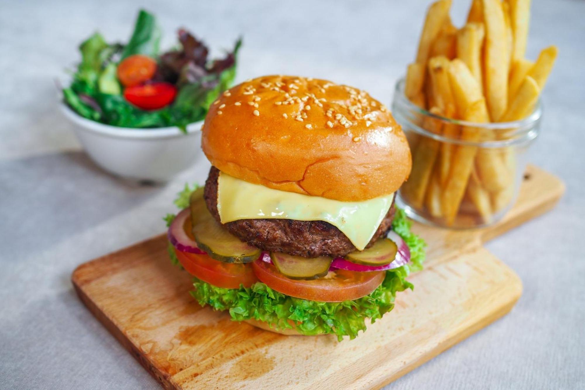 Crowne Plaza Changi Airport By Ihg Сінгапур Екстер'єр фото The photo shows a delicious looking hamburger with a sesame seed bun. The burger is layered with a beef patty, melted cheese, lettuce, tomato slices, pickles, and red onion. It is presented on a wooden cutting board. Next to the burger, there is a sm