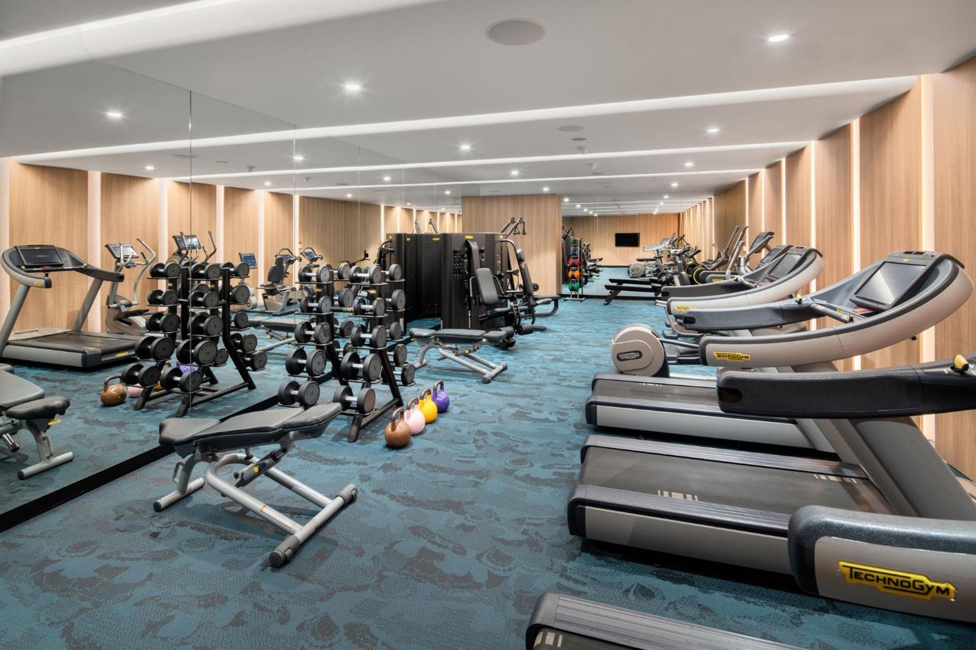Crowne Plaza Changi Airport By Ihg Сінгапур Екстер'єр фото The photo shows a modern gym interior. It features various fitness equipment, including multiple treadmills, weightlifting machines, and a set of free weights on racks. The flooring has a blue carpet, and the walls are made of wood paneling, giving t
